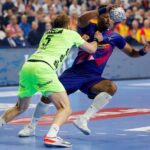 Two handball players compete intensely on the court, one in a neon green uniform defending against another in a blue and red uniform who is holding the ball above his head.