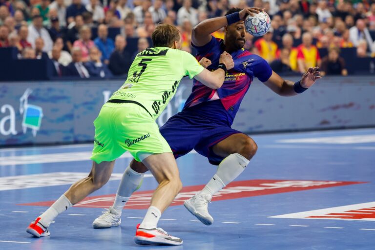 Two handball players compete intensely on the court, one in a neon green uniform defending against another in a blue and red uniform who is holding the ball above his head.